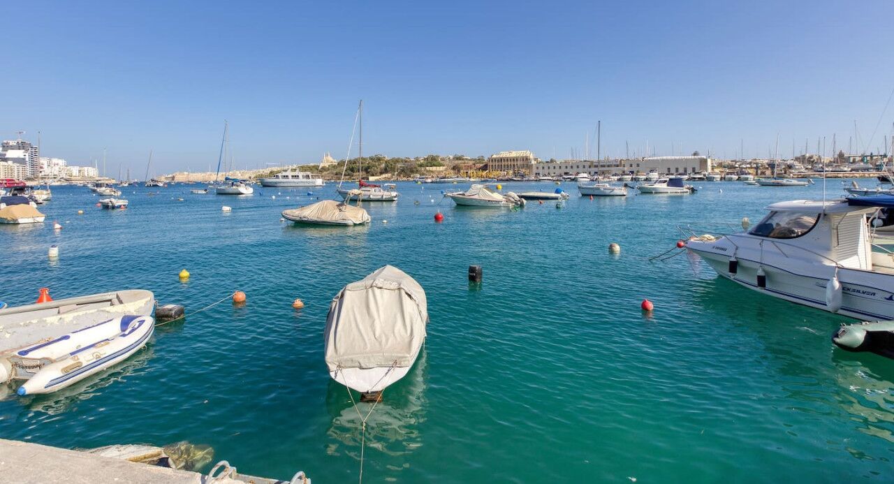 A marina with various boats docked, a clear sky, and buildings in the background.