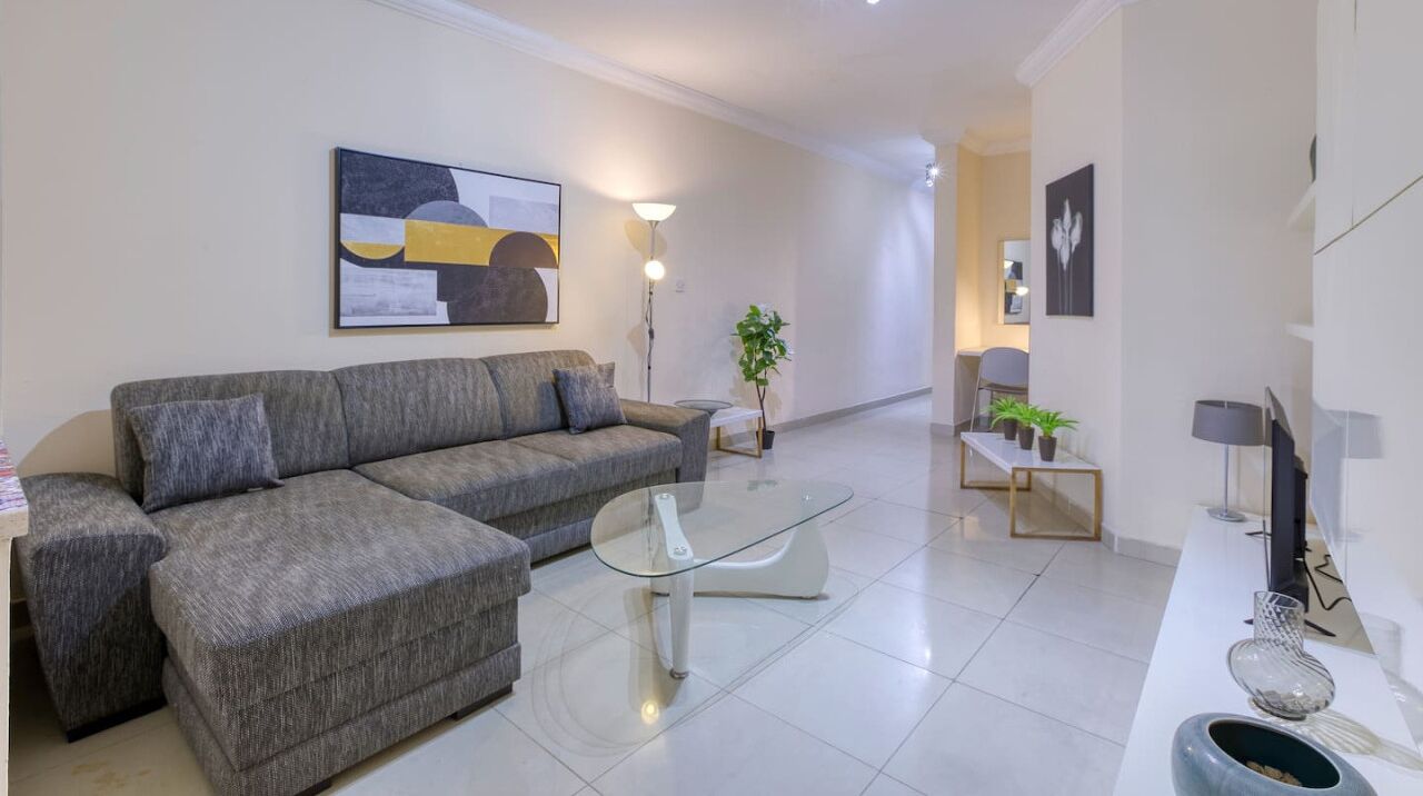 Una sala de estar moderna con un sofá gris, una mesa de centro de vidrio y un aparador blanco con objetos decorativos.
