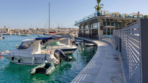 A marina with boats docked, a glass-roofed building with outdoor seating, and a clear view of the water.