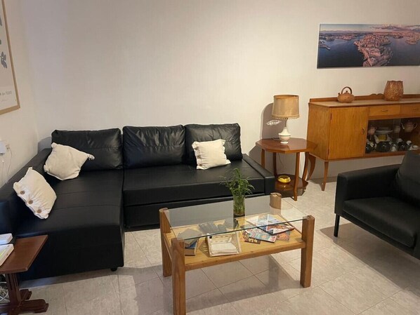 A living room with a black leather sofa, a glass coffee table, a wooden side table, and a cabinet with kitchenware.