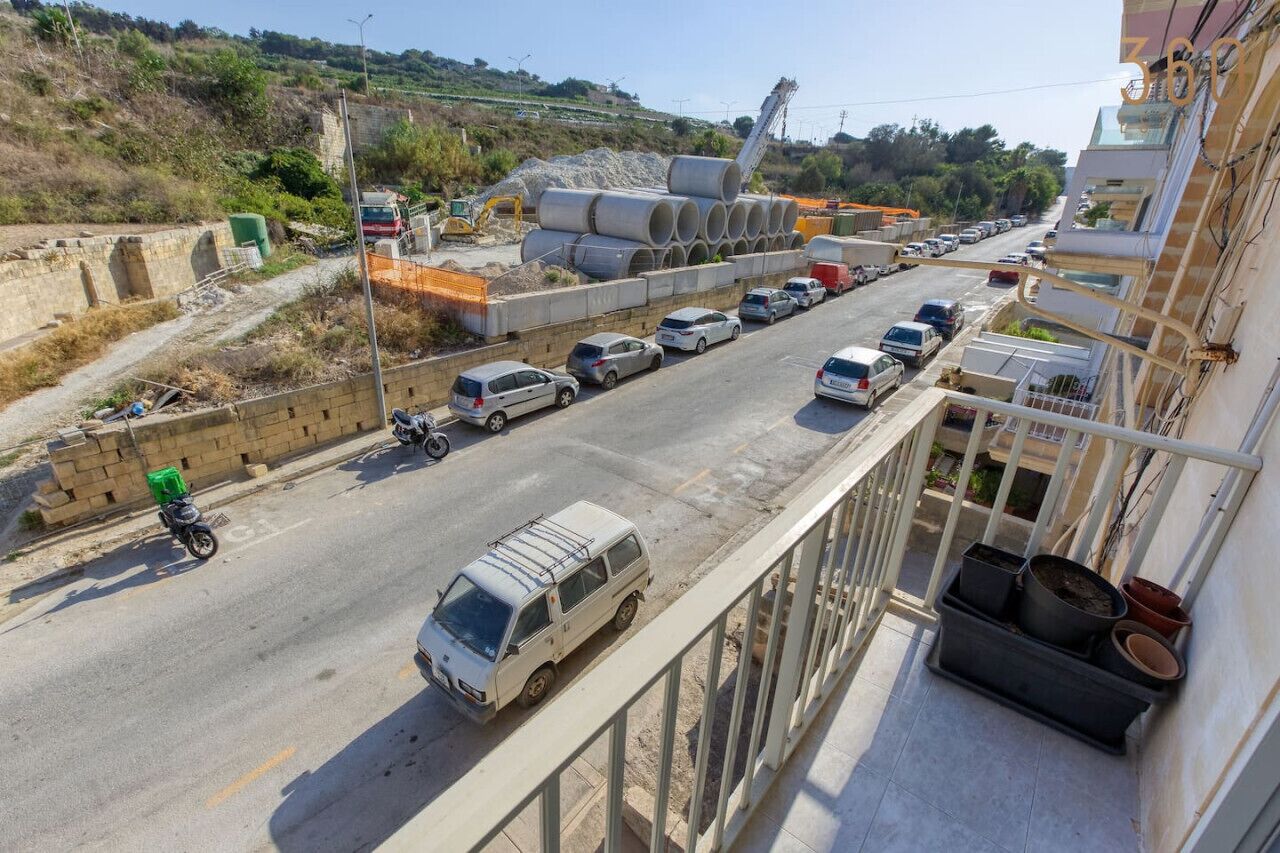 Un chantier de construction avec des véhicules et du matériel sur une route, un fourgon garé sur le bord, et un balcon orné de plantes en pot.