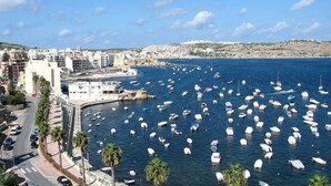 A coastal cityscape with a marina filled with boats, buildings along the waterfront, and a clear sky.