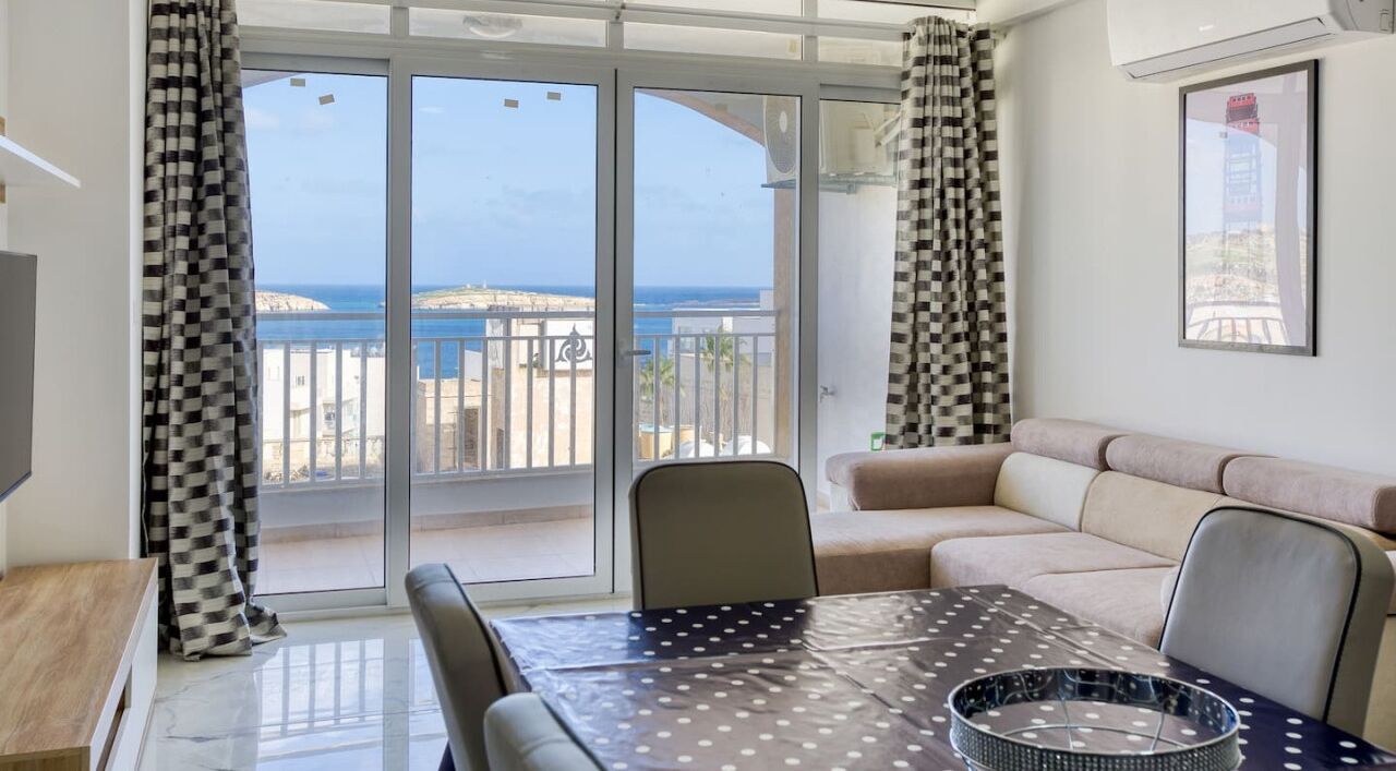 Un salon avec un canapé, une table à manger recouverte d’une nappe à pois, et une vue sur la mer par de grandes portes vitrées.