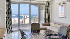 A living room with a sofa, a dining table with a polka dot tablecloth, and a view of the sea through large glass doors.