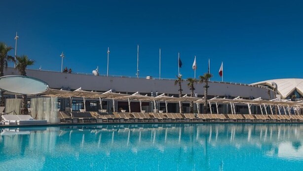 A poolside area with lounge chairs and flags, a clear blue sky, and a building with a curved roof.
