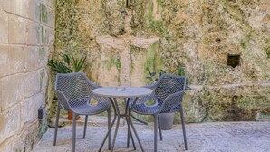 Un petit espace extérieur aménagé avec une table ronde et deux chaises, adossé à un mur à la texture particulière, percé d’une petite fenêtre.