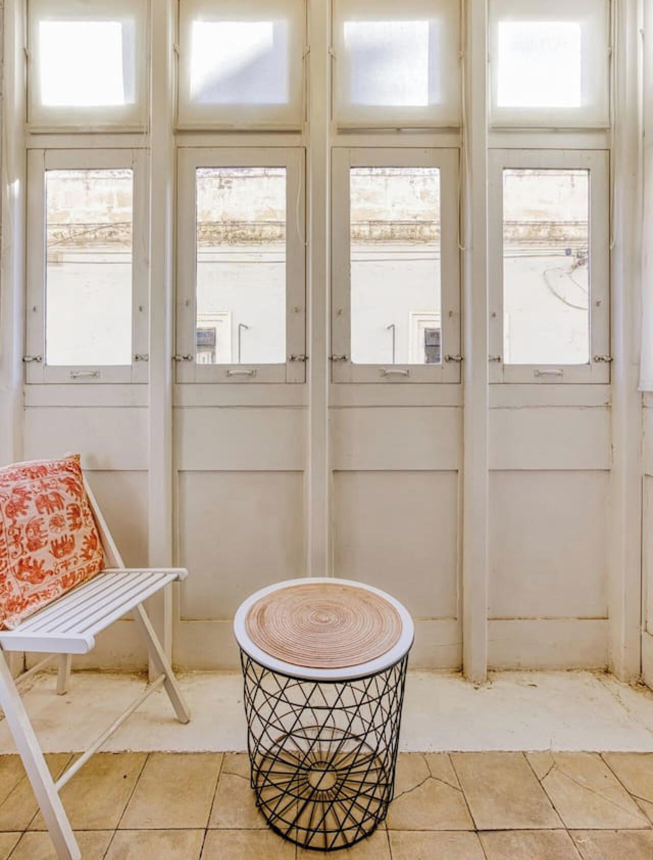 Une pièce avec une chaise blanche et une table ronde dotée d’un piétement en osier, une fenêtre donnant sur un immeuble, et une porte avec une poignée.
