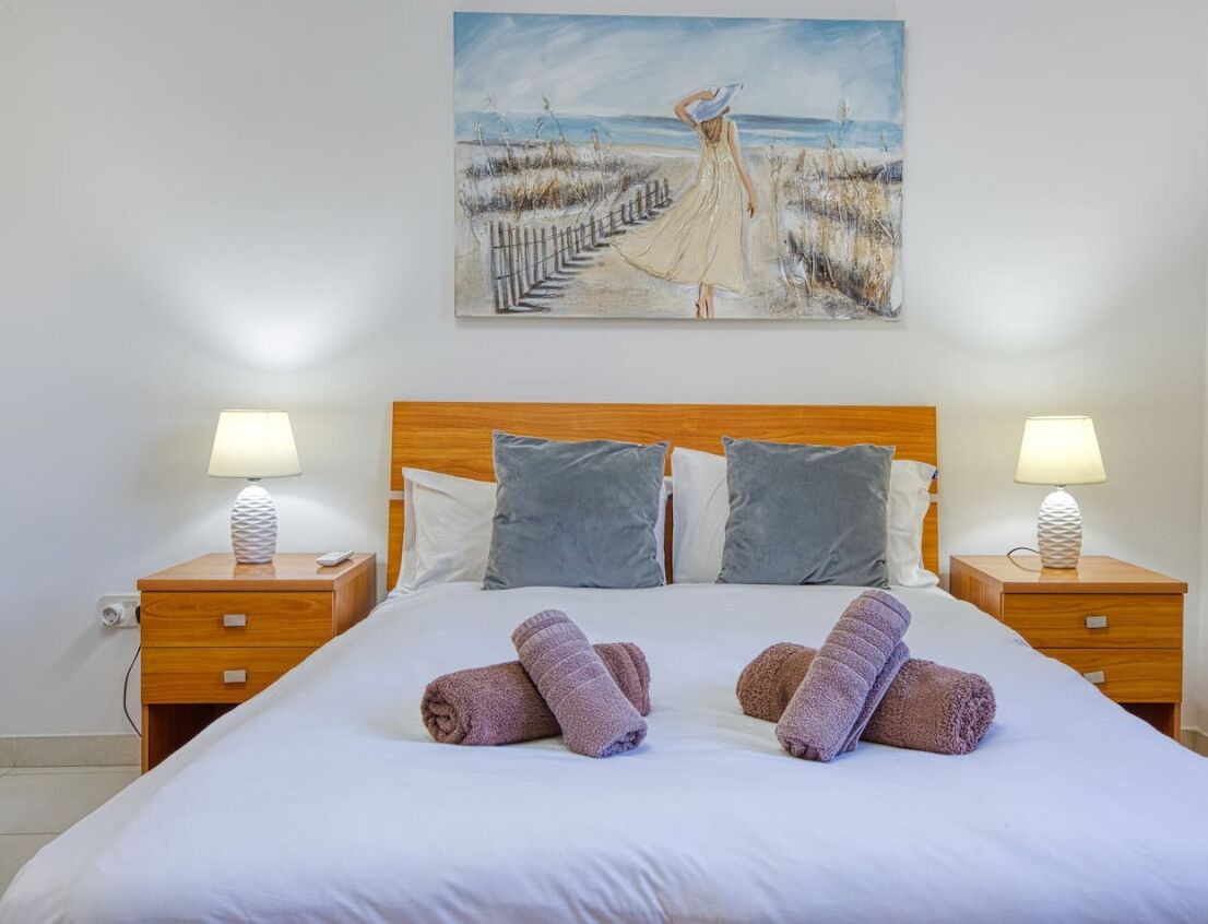 Una cama bien hecha con dos almohadas, dos mesitas de noche con lámparas, y un cuadro de una playa en la pared.