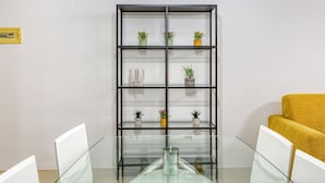 Un salón de conferencias con una mesa de vidrio, sillas blancas, un sofá amarillo y un estante negro con plantas en macetas.