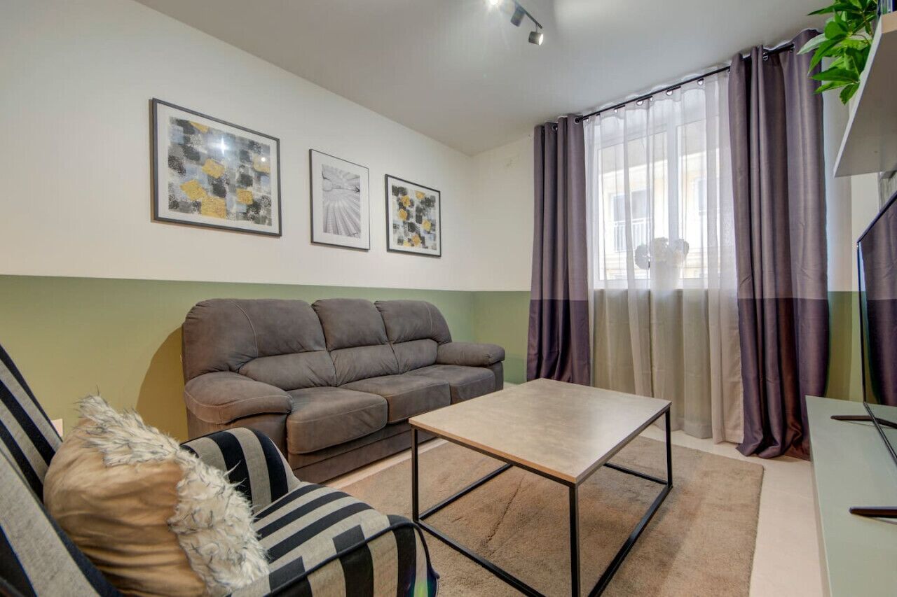 Un salon avec un canapé gris, un fauteuil à rayures, une petite table et une fenêtre avec des rideaux.