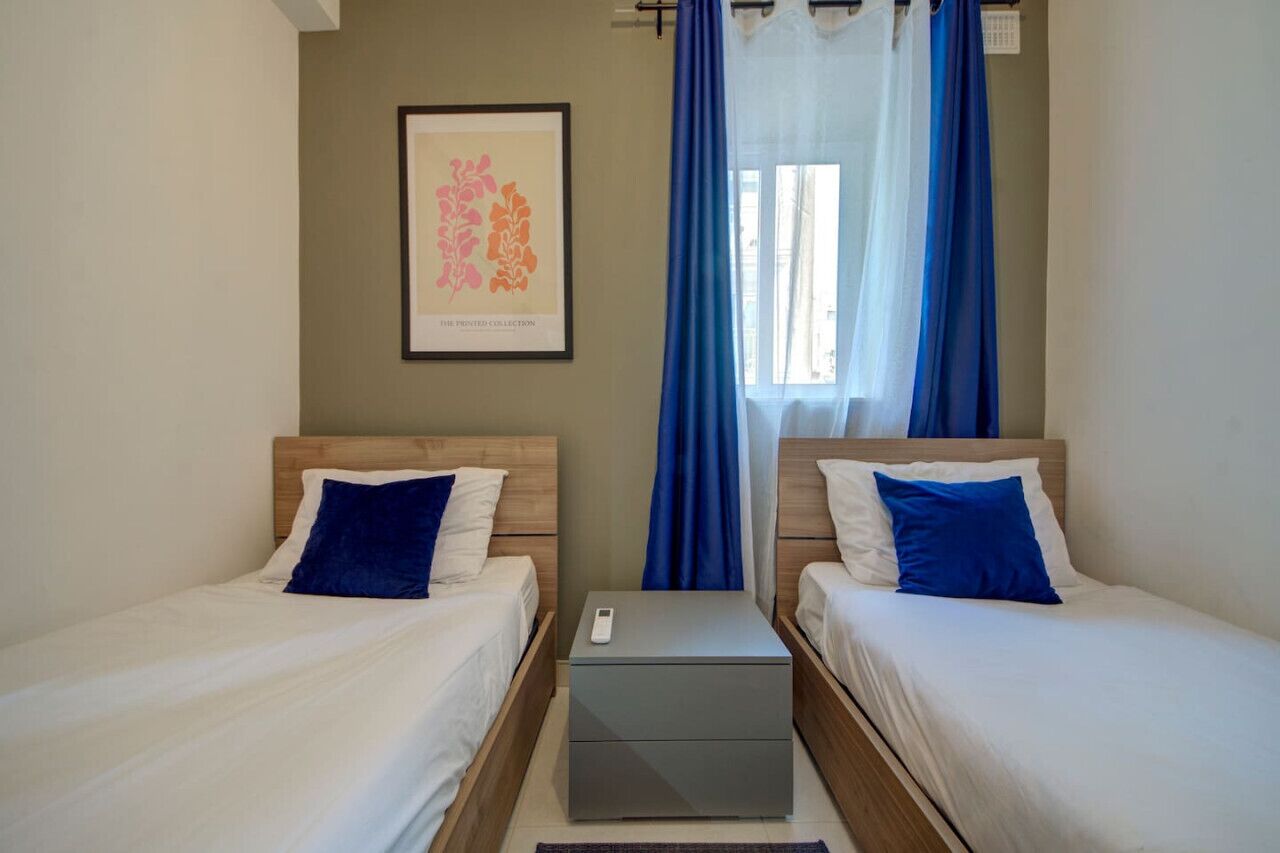 Habitación doble con dos camas, una mesita de noche con teléfono, una obra de arte enmarcada en la pared y una ventana con cortinas azules.