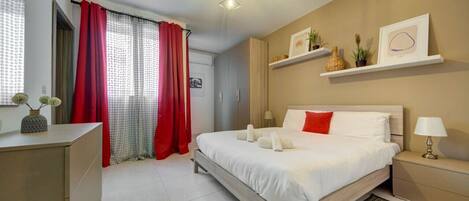 Habitación de hotel moderna con una cama grande, una mesita de noche con lámpara, un escritorio pequeño y un estante con objetos decorativos.