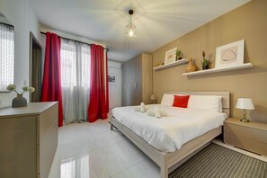 A modern hotel room with a large bed, a bedside table with a lamp, a small desk, and a shelf with decorative items. - Modern 2BR home with modern amenities in Gzira (Il-Gżira)