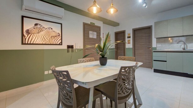 Un coin repas avec une table ronde, des chaises et un luminaire suspendu. Un tableau représentant un zèbre, encadré, est accroché au mur, et un pot de fleurs est posé sur la table.
