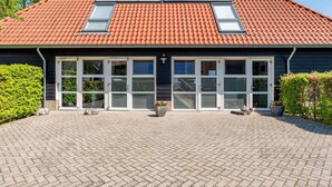 Ein Gebäude mit rotem Ziegeldach, großen Fenstern und einem gepflasterten Eingangsbereich mit Kübelpflanzen.