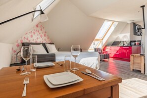 Mesa de jantar posta com pratos, talheres e copos de vinho em um quarto com teto inclinado, cama com roupa de cama florida e área de cozinha vermelha.