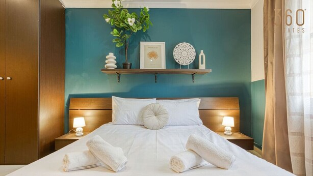 A neatly made bed with white linens and two rolled towels at the foot of the bed. There are two bedside lamps on wooden nightstands, a wall-mounted shelf with decorative items, and a teal-colored wall. - Stylish 1BR Retreat w/ Balcony in Prime St Julian’s (St. Julian`s)