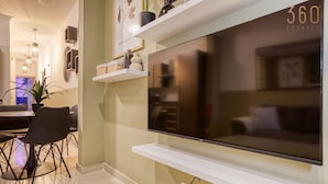 A modern living room with a large flat-screen TV mounted on a white wall, a dining area with a table and chairs, and decorative shelves with various items. - Stylish 1BR Retreat w/ Balcony in Prime St Julian’s (St. Julian`s)