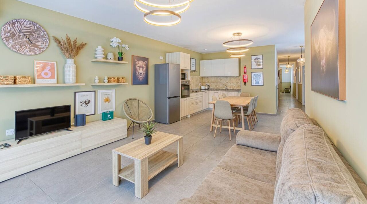 A modern living room with a sofa, a coffee table, a TV, and a fireplace. There is a kitchen area with a refrigerator, stove, and dining table.