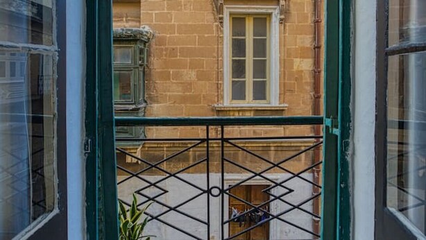 Vista desde una puerta de balcón de un edificio con fachada de piedra, una ventana y una reja metálica.