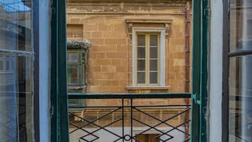 Une vue à travers la porte d’un balcon, donnant sur un immeuble à la façade en pierre, avec une fenêtre et un portail en métal.