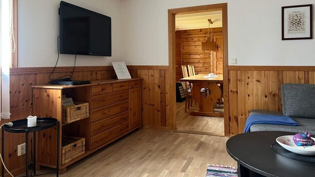 A room with wooden paneling, a flat-screen TV mounted on the wall, a dining table with chairs, a sofa, and a small table with a plate of food. - 6 Person Holiday Home in Bredaryd (Bredaryd)