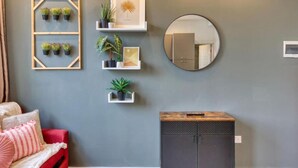 A room with a grey wall, a round mirror, and three white shelves with plants and pictures. There is a red sofa with pink pillows and a small cabinet with a wooden top.