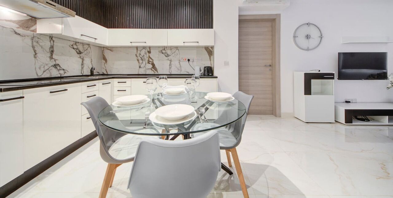 A modern kitchen with a glass dining table, white cabinets, and a marble backsplash.