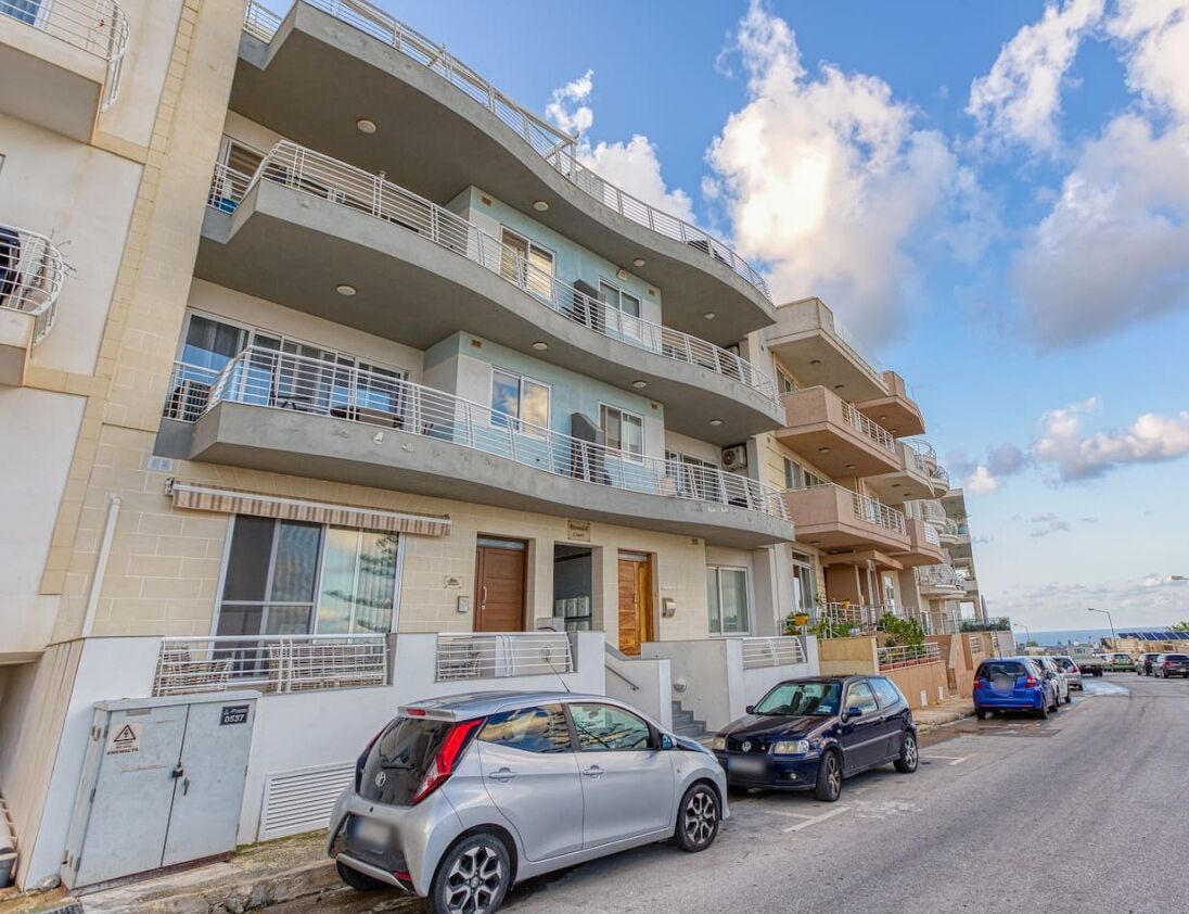 Un edificio residencial de varios pisos con balcones, un área de estacionamiento y un cielo despejado.
