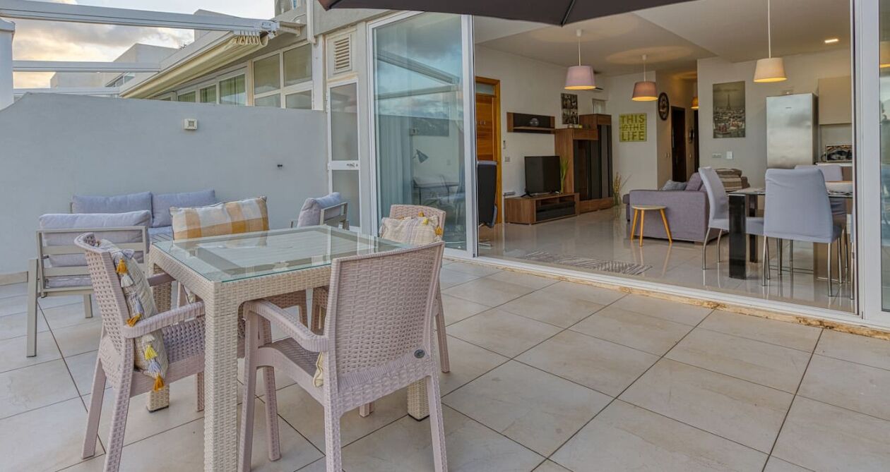 A modern outdoor dining area with wicker furniture and a glass table, leading to a glass door into a living space with a TV and dining table.