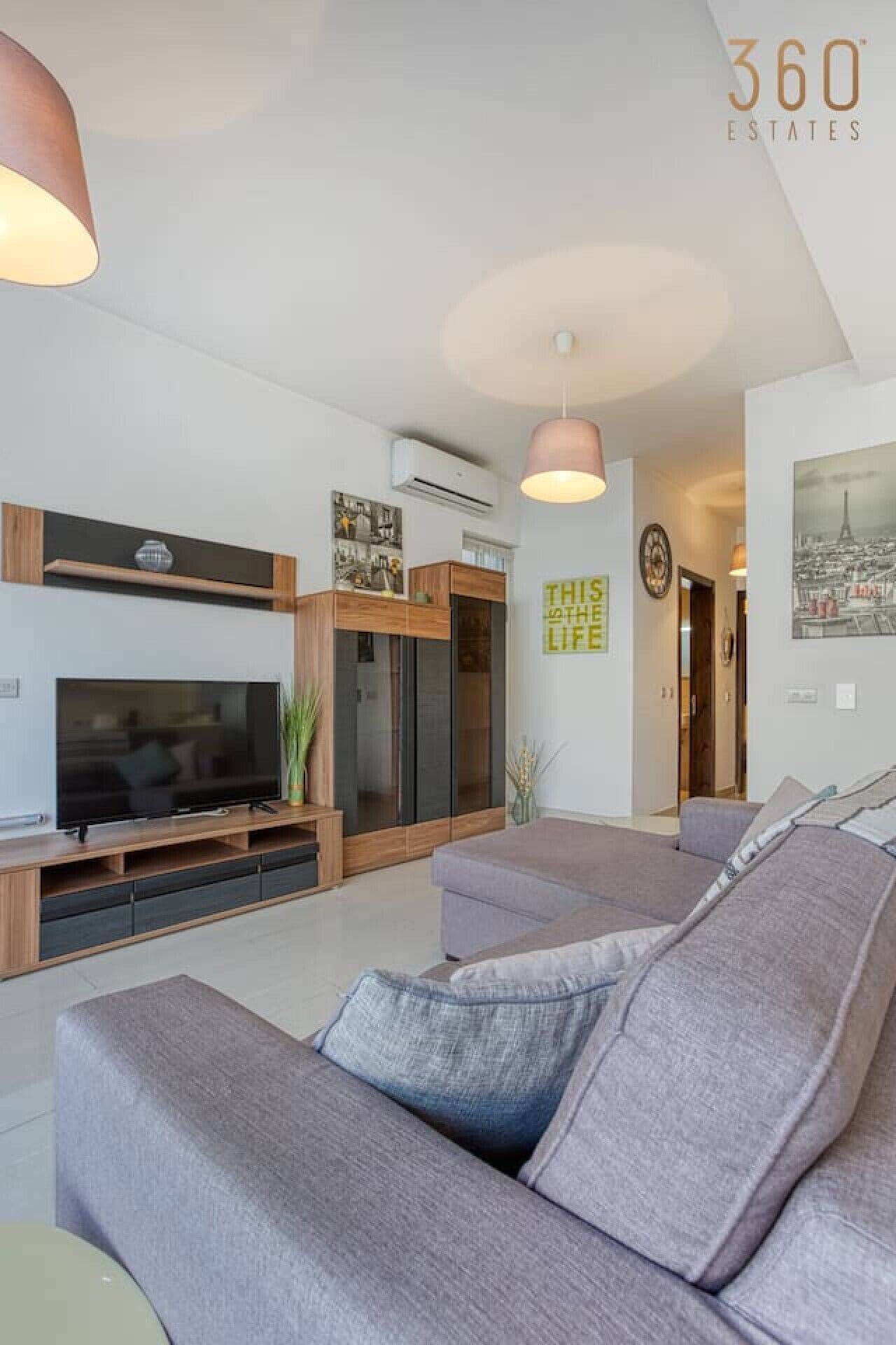 A modern living room with a grey sofa, a flat-screen TV, wooden cabinets, and a pendant light.