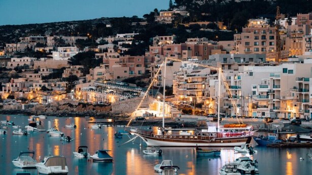 A coastal town with a marina filled with boats, buildings with balconies, and a prominent ship docked at the harbor.