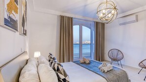 A hotel room with a large bed, a view of the sea, a chandelier, and a chair.