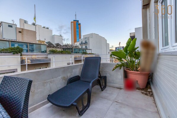 A balcony with a chair and a potted plant, overlooking a cityscape with modern buildings.