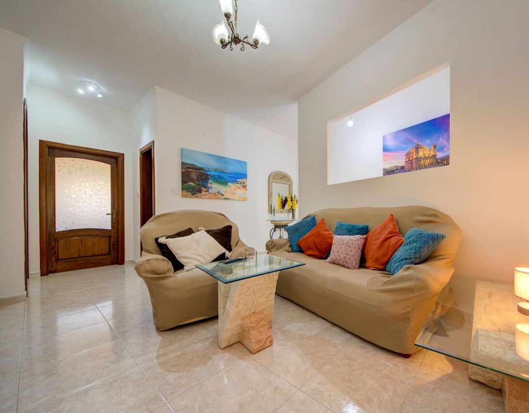 A living room with a beige sofa, a glass coffee table, and a chandelier. There are paintings on the walls and a door with frosted glass.