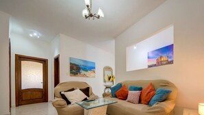 A living room with a beige sofa, a glass coffee table, and a chandelier. There are paintings on the walls and a door with frosted glass. - Traditional Maltese Escape with Rooftop Terrace! (Ħad-Dingli)