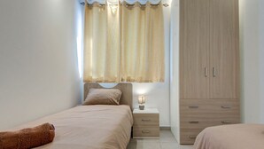 A bedroom with two beds, a nightstand with a lamp, a wardrobe, and a window with curtains. - Modern 3BR home with Balcony in Central Msida (L-Imsida)