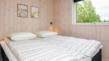 A bedroom with a wooden paneled wall, a bed with white bedding, two framed pictures, and a window with a view of trees.