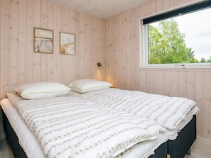 A bedroom with a wooden paneled wall, a bed with white bedding, two framed pictures, and a window with a view of trees.