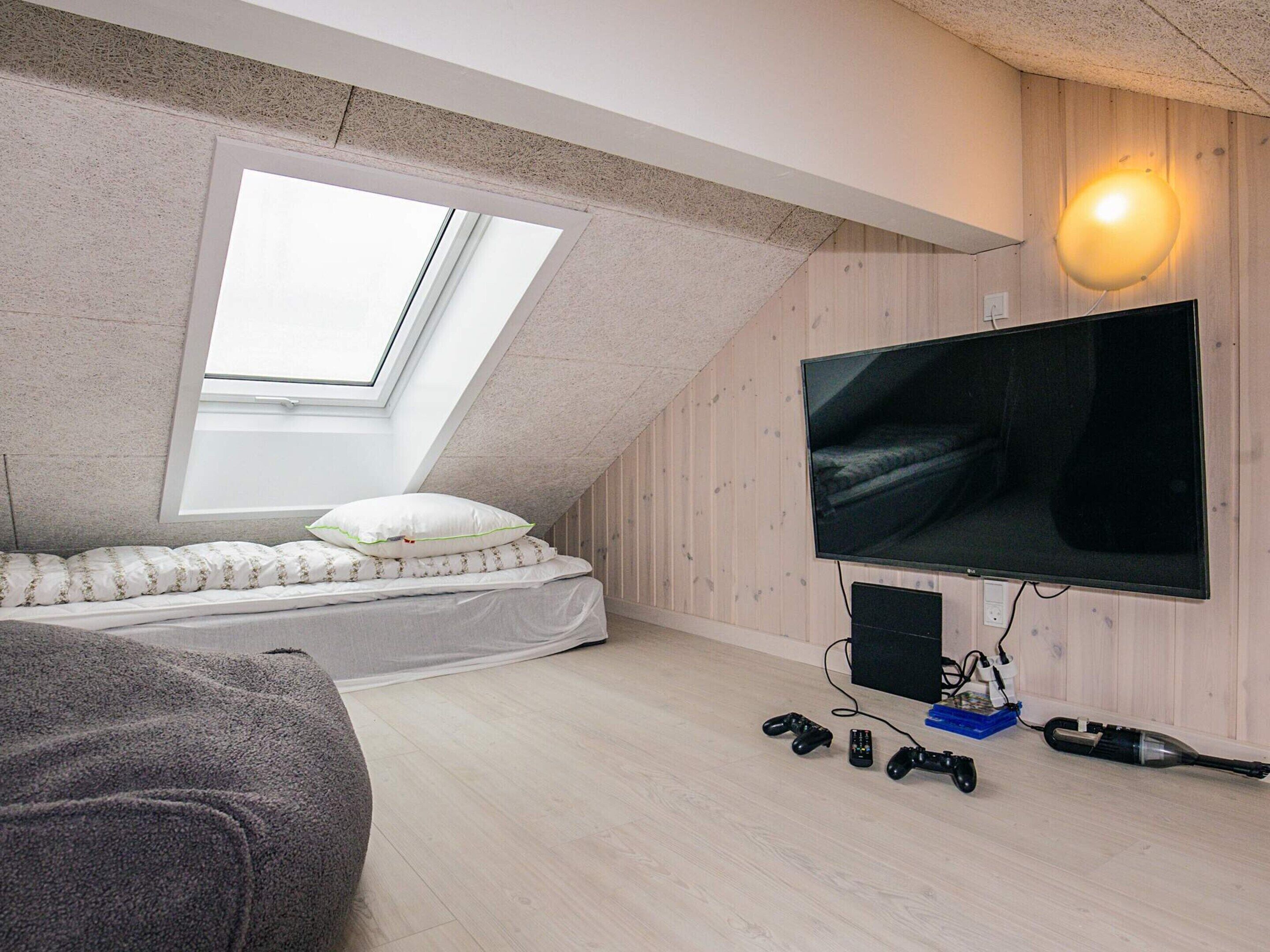 Ein Zimmer mit schräg angelegter Decke, einem Bett mit weißer Bettwäsche, einem an der Wand montierten großen Fernseher und einer Deckenleuchte.