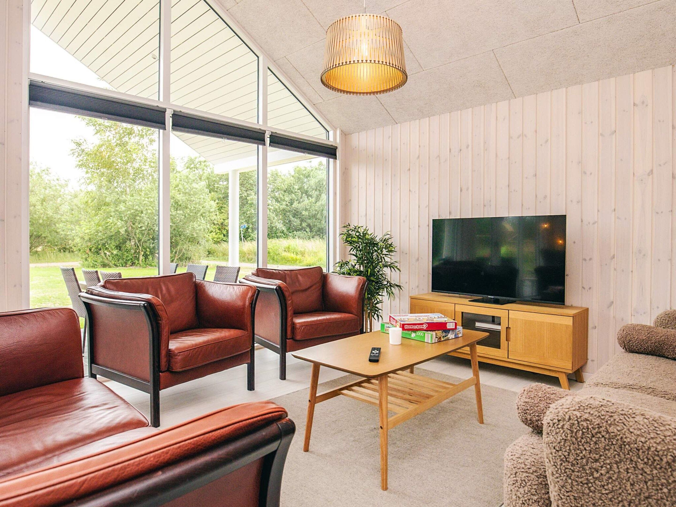 Ett modernt vardagsrum med lädersoffor, ett soffbord i trä, en tv placerad på ett träskåp och stora fönster med utsikt över grönområden.