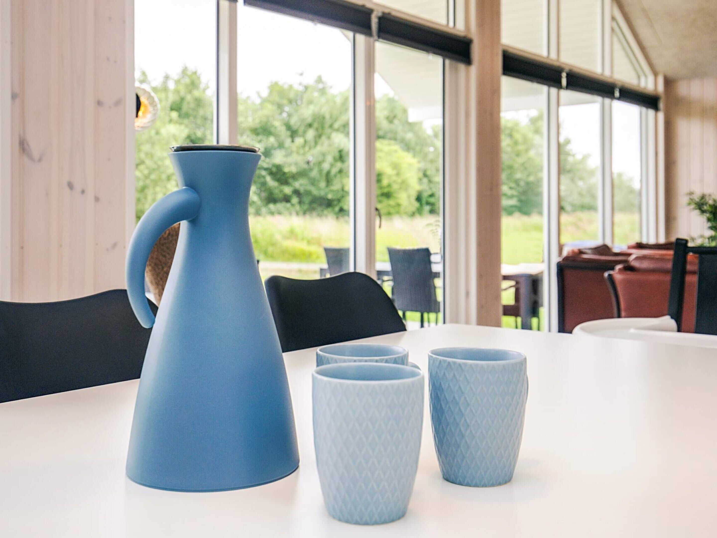 A table with a blue pitcher and two patterned cups, set in a room with large windows and outdoor views.