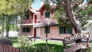 Un edificio de dos pisos de ladrillo con techo de paja, un letrero que dice 'NONQUEN DUPLEX CABANAS', y un pequeño espacio de estar al aire libre.
