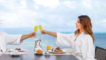 A couple toasting with champagne glasses at a table with breakfast, overlooking a scenic ocean view.