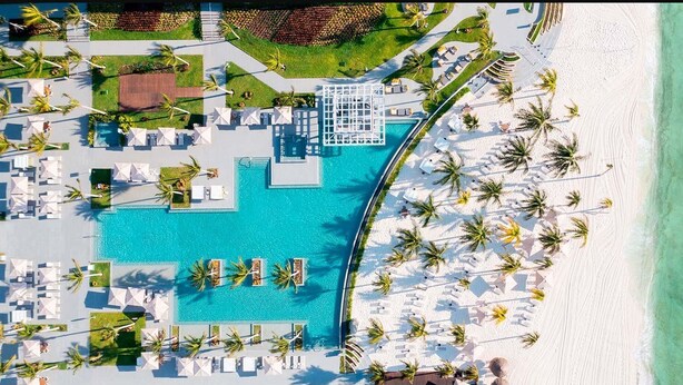 Vista aérea de un complejo turístico con una piscina central, varias zonas de descanso con sombrillas y una playa con palmeras.
