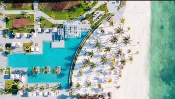 An aerial view of a resort with a central swimming pool, multiple lounge areas with umbrellas, and a beach area with palm trees.