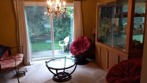 Ein Zimmer mit Glastür, die zu einem Garten führt, einem runden Tisch, einem roten Sessel und einem Schrank mit Glastüren.