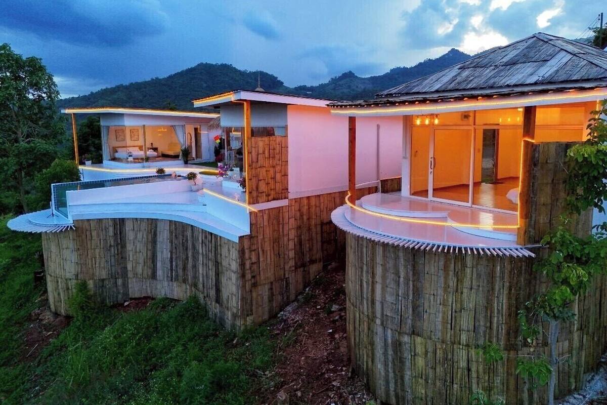 A wooden structure with a flat roof and a balcony, surrounded by greenery and hills.