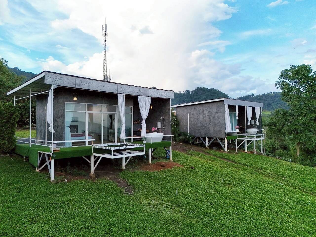 Dos cabañas modernas con paredes de vidrio y cortinas blancas, ubicadas en una colina cubierta de pasto bajo un cielo despejado.