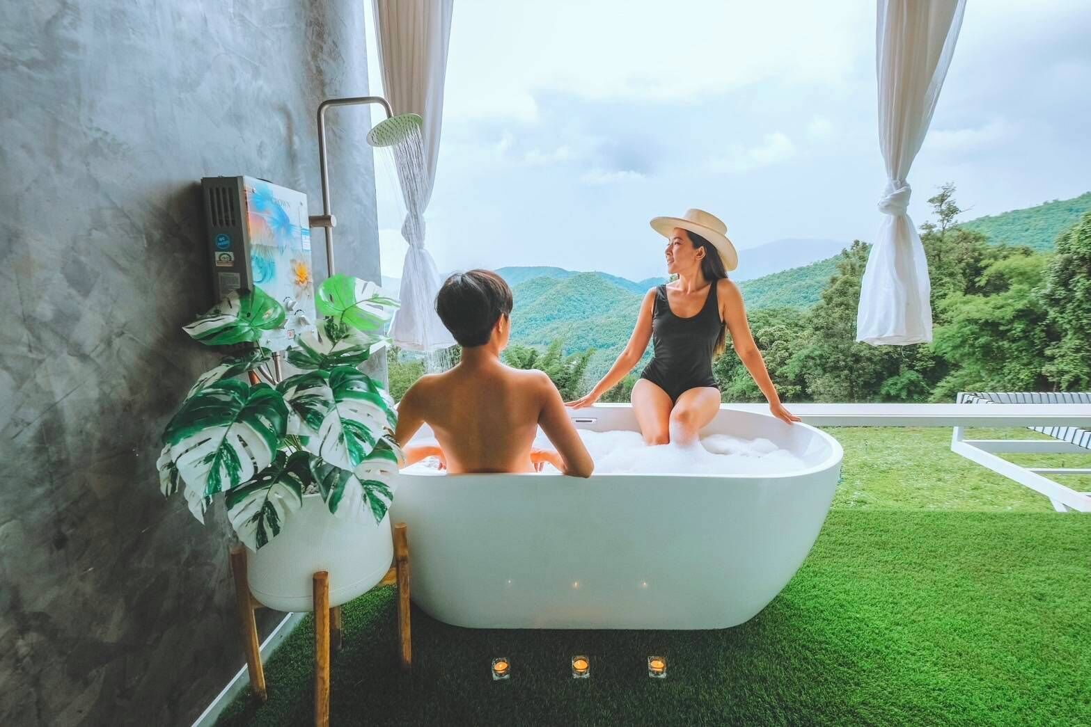 A bathtub with two people, a large potted plant, and a view of green hills.